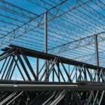 Looking up at steel joists under a blue sky before the roof has been laid.